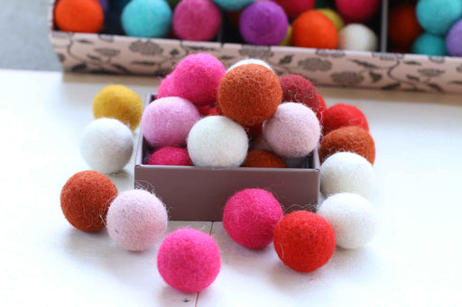 A collection of colorful felt pom-poms in various shades of pink, orange, red, white, yellow, and purple, displayed in a small box with more of the same in the background.