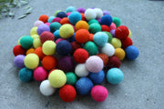 A pile of colorful felt pom-poms in various colors including red, blue, green, yellow, pink, orange, purple, and white, arranged on a gray concrete surface.