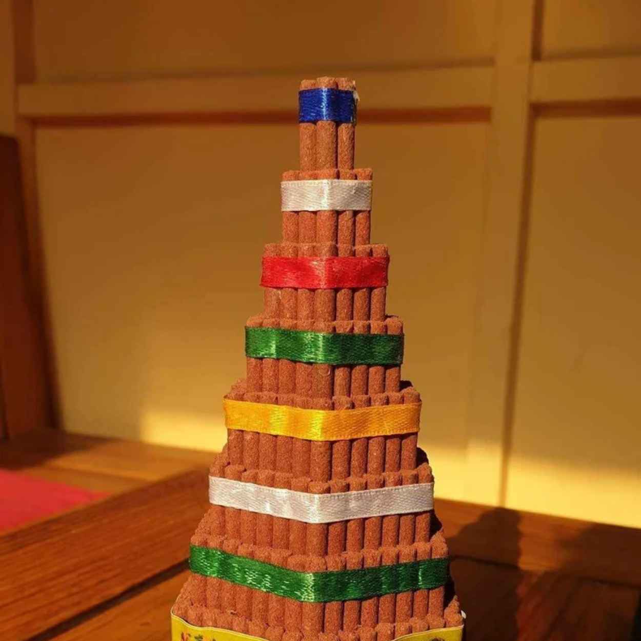 A decorative miniature pagoda-shaped tower made from stacked brown cylindrical stick incense , accented with multiple colored ribbons in blue, silver, red, green, yellow, and white, displayed on a wooden surface against a warm-toned indoor background.