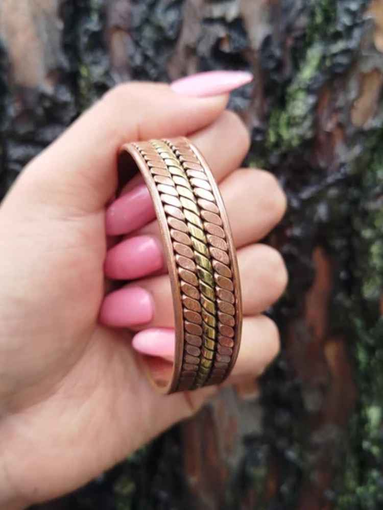 A close-up of a hand with pink manicured nails holding a wide, textured metallic bangle bracelet. The bracelet has a rose gold finish with a central gold accent stripe.