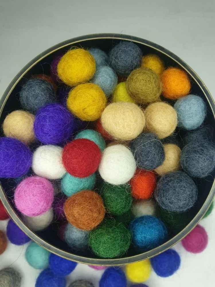 A top-down view of a round metal tin filled with colorful wool felt balls in various colors including yellow, blue, purple, red, white, green, orange, and pink, set against a neutral light gray background.