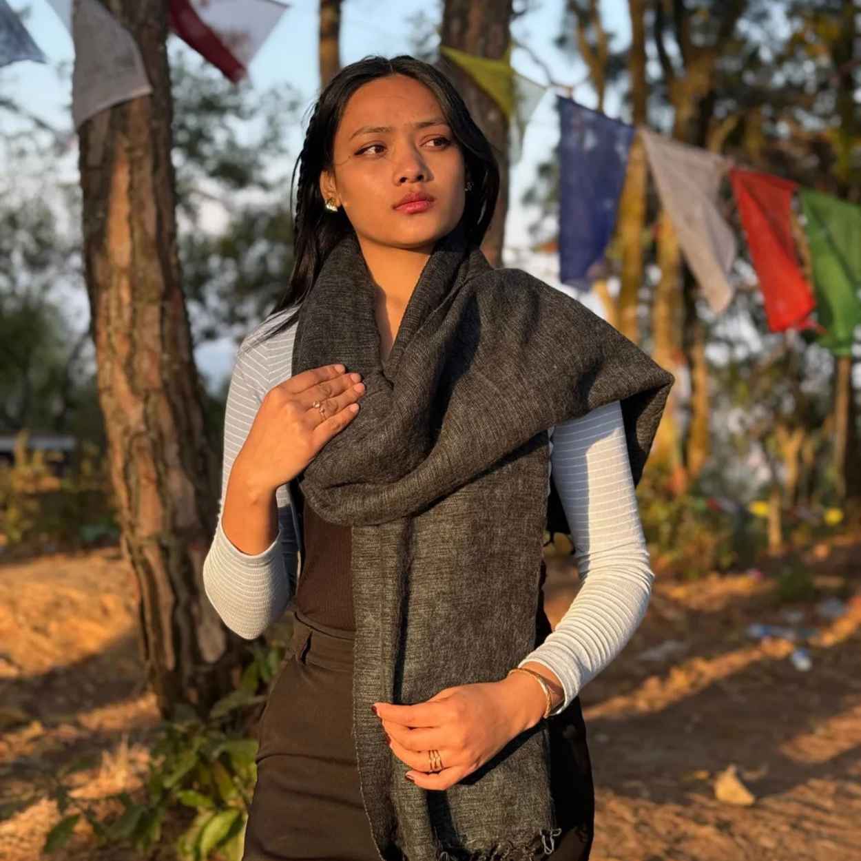 A woman wearing a dark gray textured knit scarf wrapped around her shoulders, standing outdoors in a wooded area with colorful prayer flags hanging in the background.